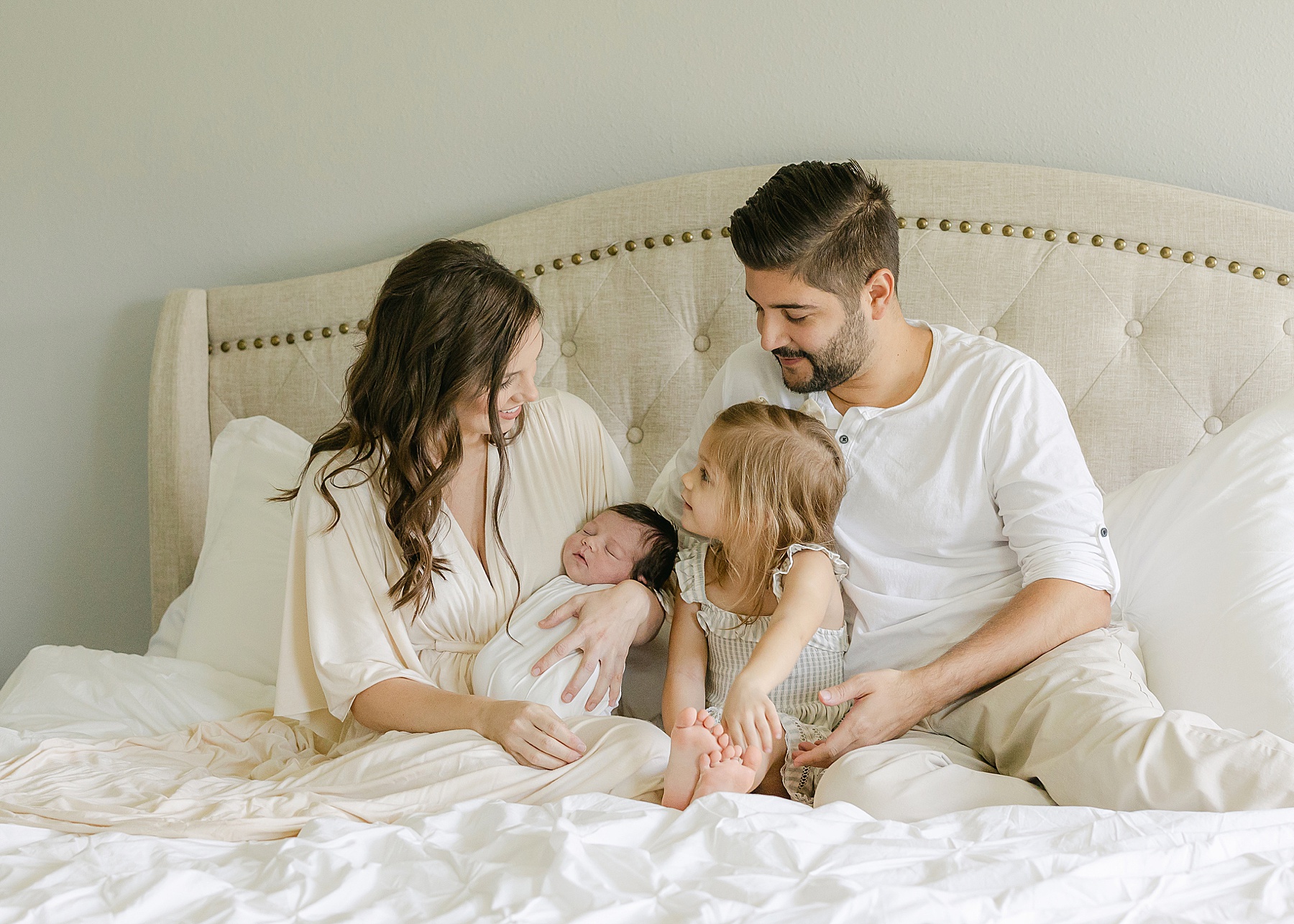 family dressed in neutral clothing sitting on bed holding baby boy