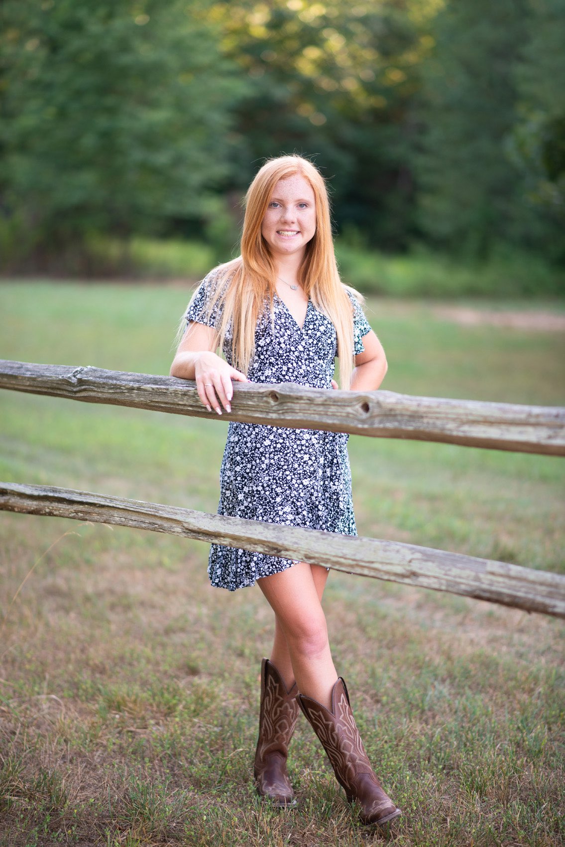 Senior girl in a print dress standing in front of a split rail fence.