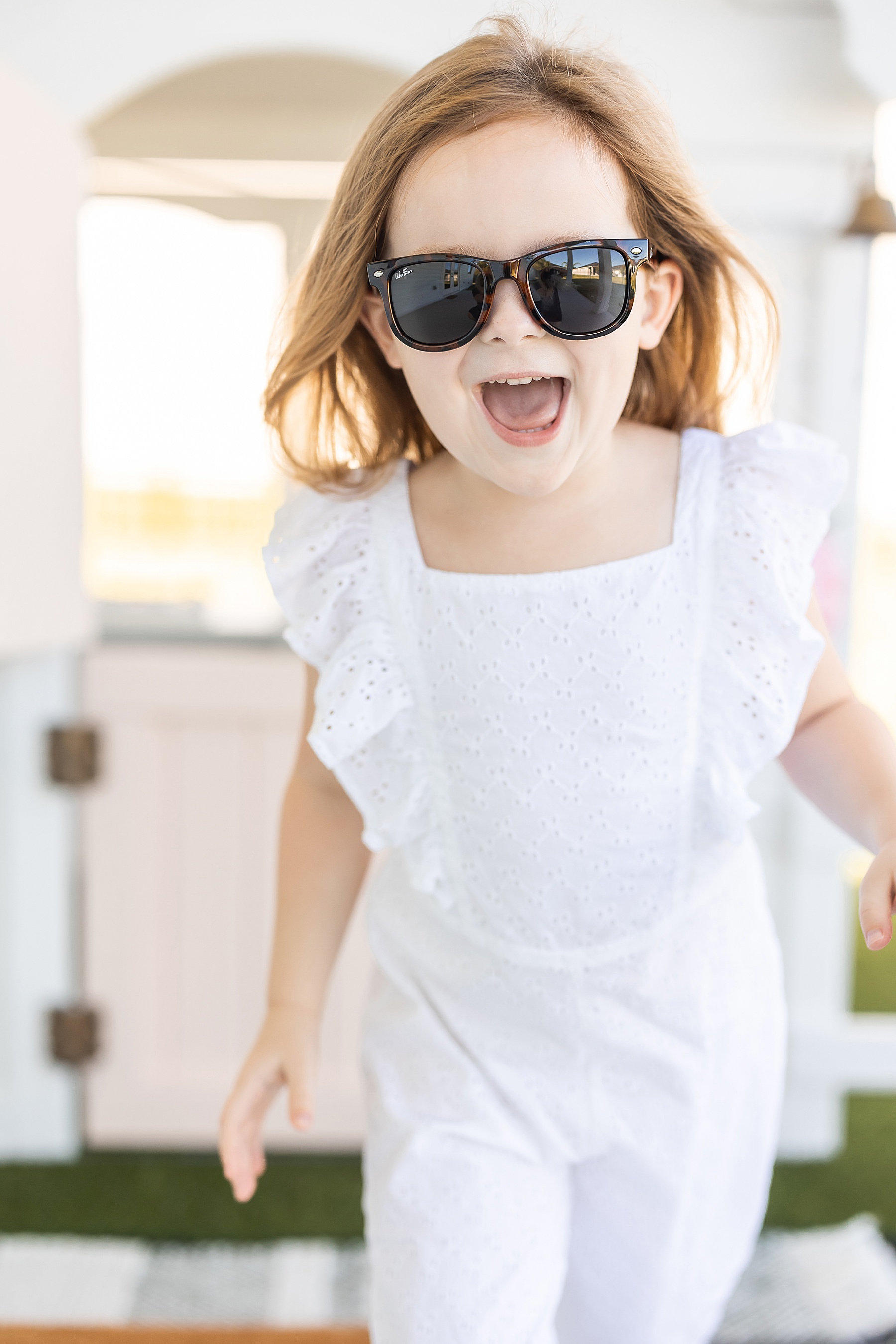 little girl in sunglasses in white jumpsuit running in front of white playhouse with pink door