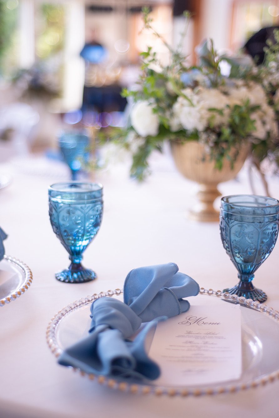 Close up of place setting for wedding reception with french blue goblets clear chargers and blue napkins.