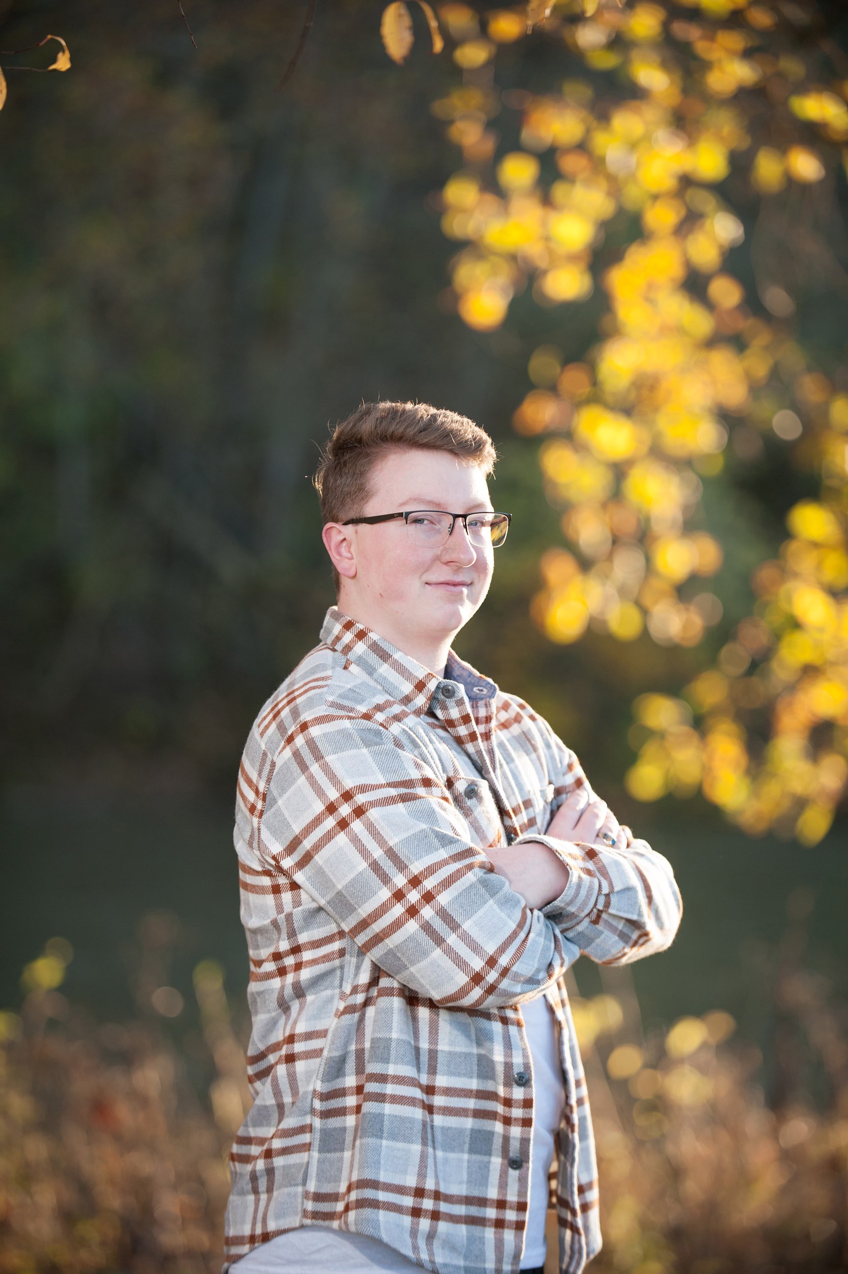 Senior guy in plaid shirt standing in front of fall foliage.