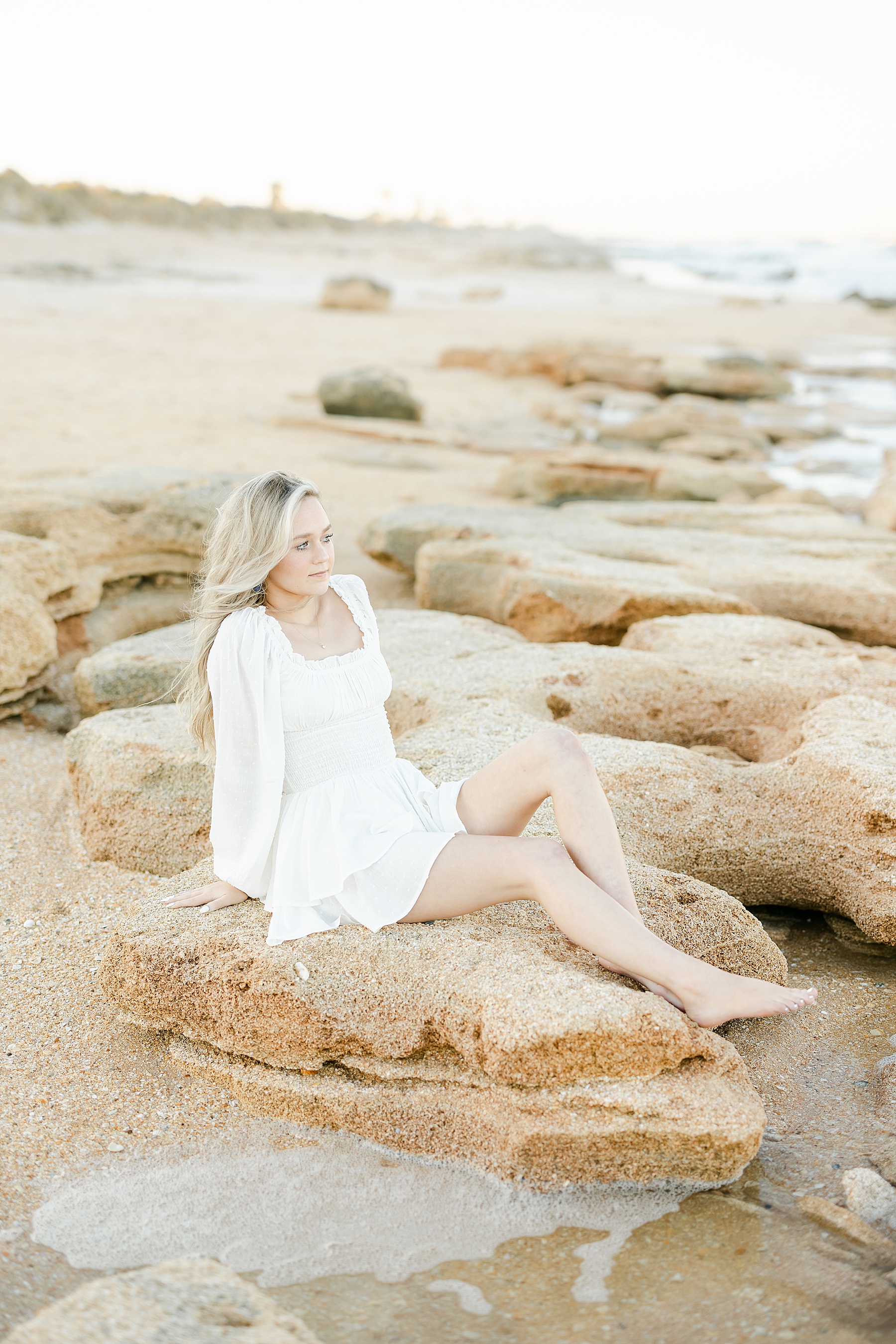 young woman sitting on rocks at the beach wearing white romper