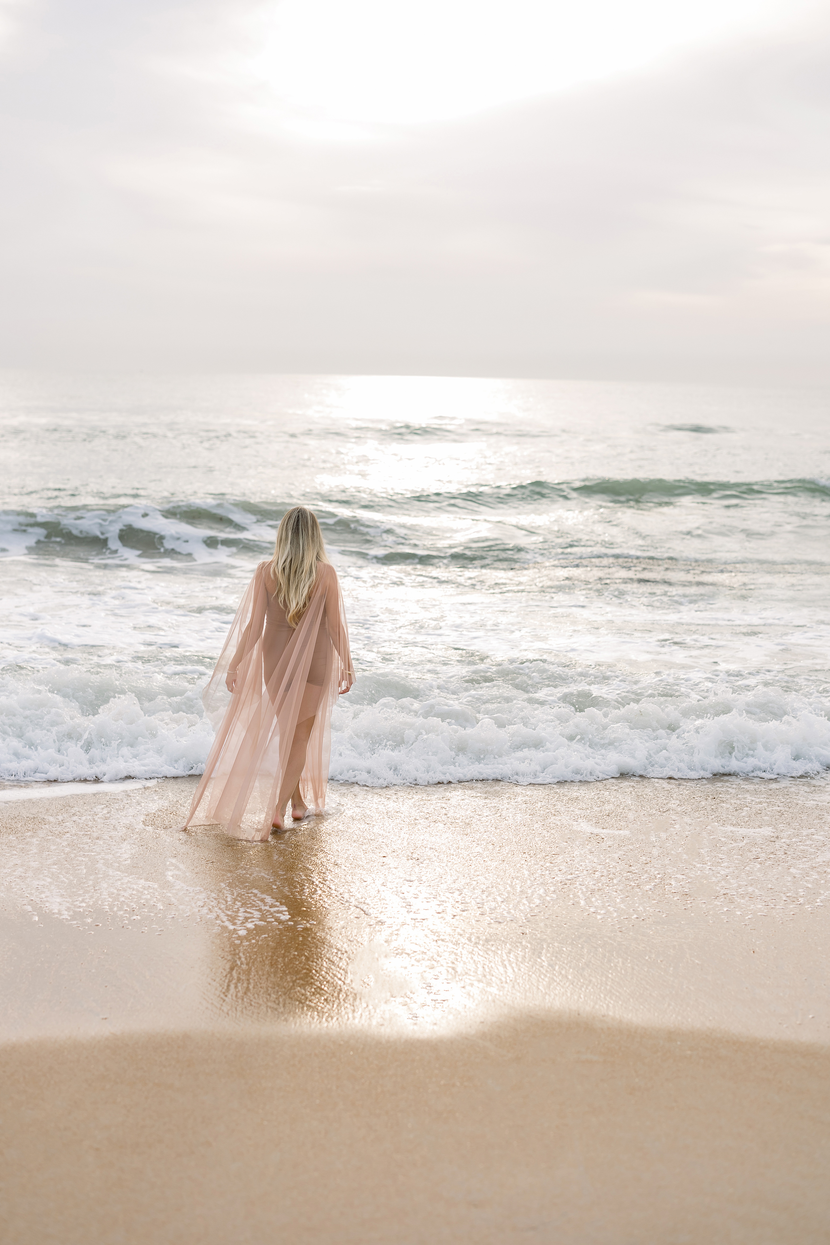 light and airy maternity portrait of blond woman standing on the sand on the beach at sunrise
