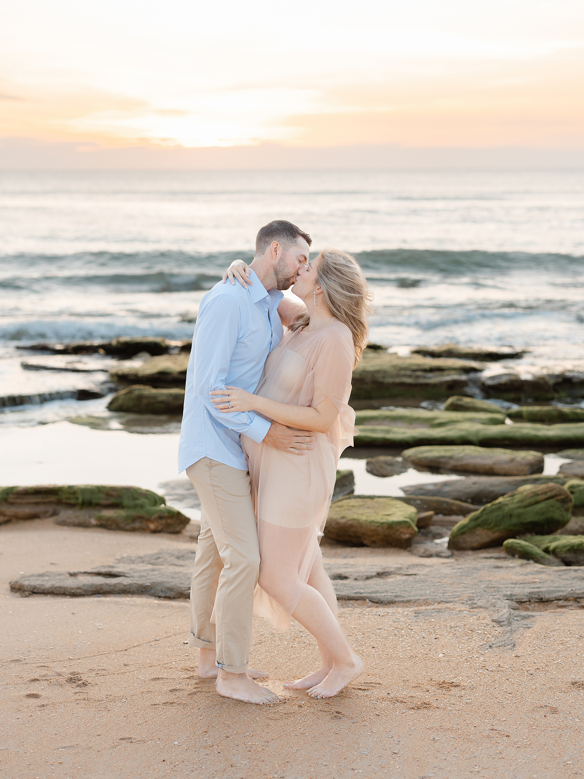 A man and pregnant woman kissing on the beach at sunrise.