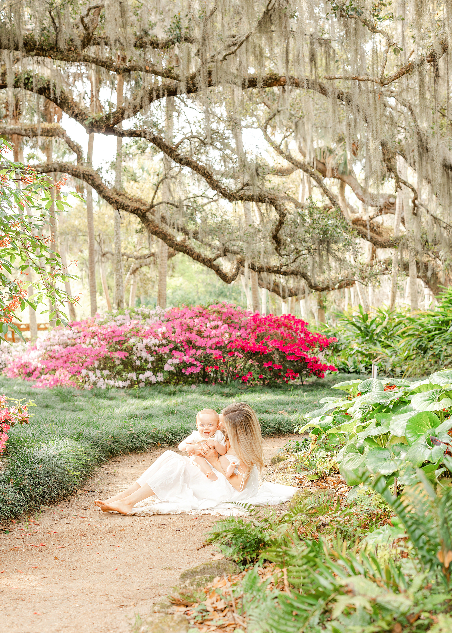 A colorful spring portrait of a woman in a white dress holding her baby boy on the ground amidst pink Azaleas at Washington Oaks.