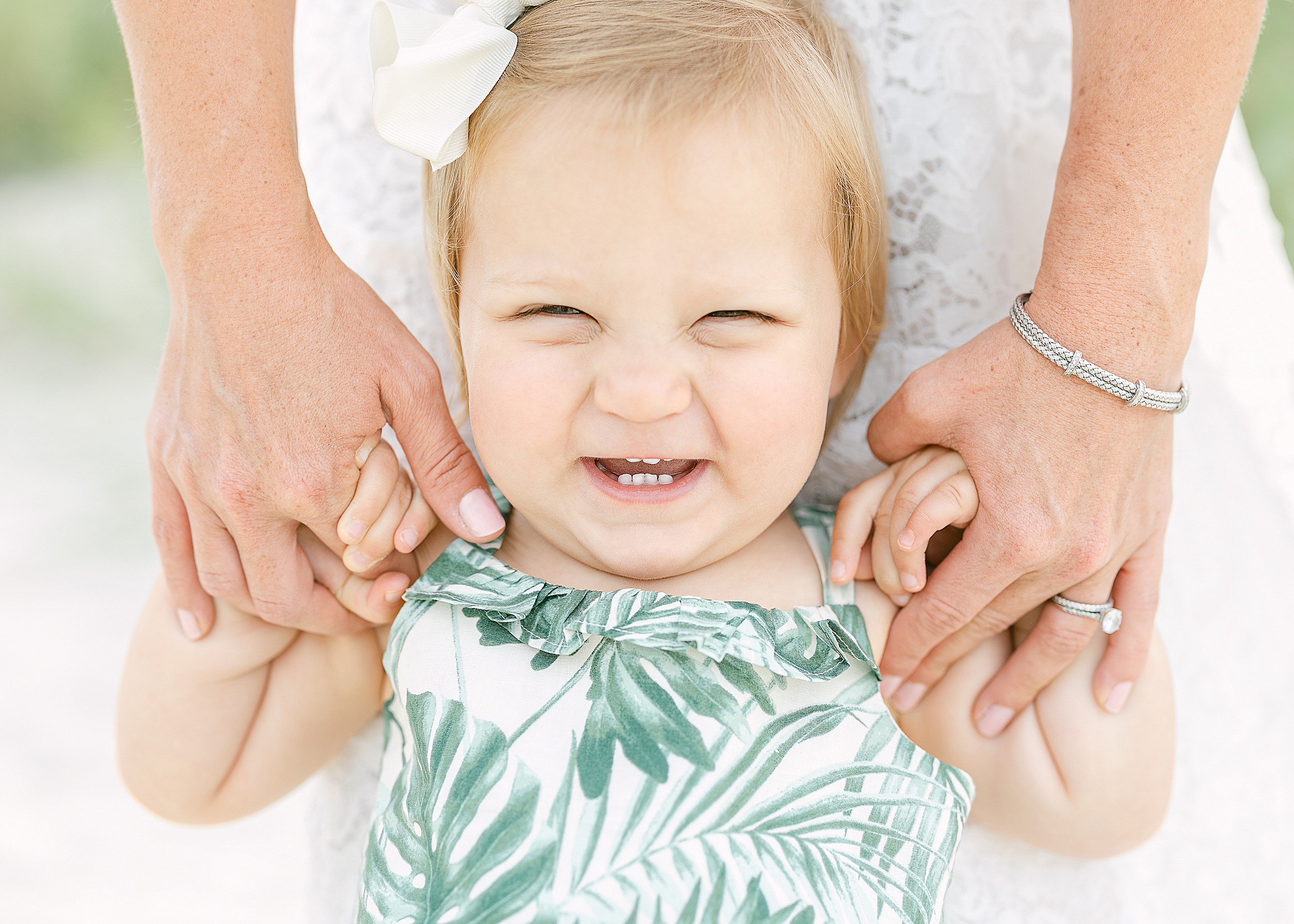 A portrait of a baby girl in a white and green palm print dress smiling big at the camera.