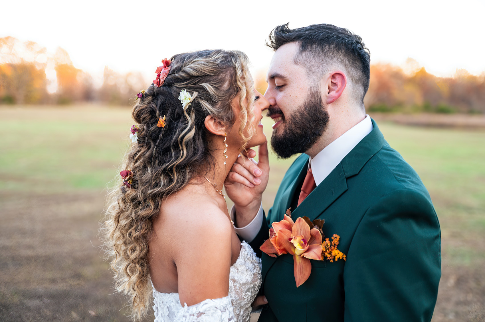 Groom in green suit and orange flower boutonniere kissing his bride at sunset. 