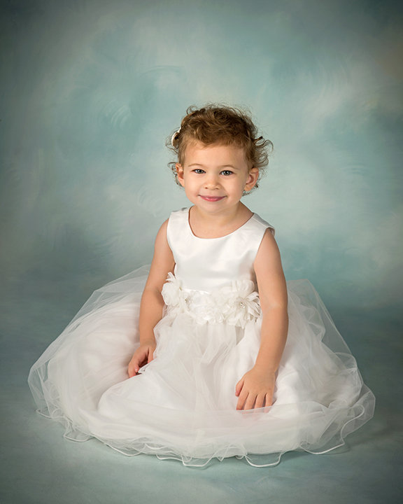 Young child in a white dress sitting on a light blue background, smiling softly at the camera.