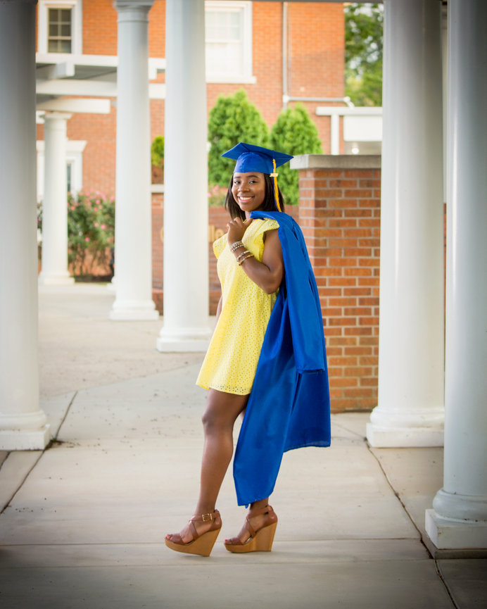 Graduate in blue cap and gown over yellow dress, standing between white columns and smiling, with brick building in background.