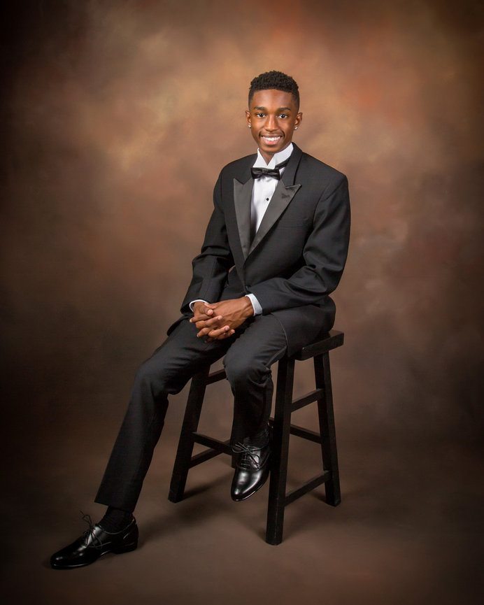 Man in a black tuxedo sitting on a stool against a soft brown background.