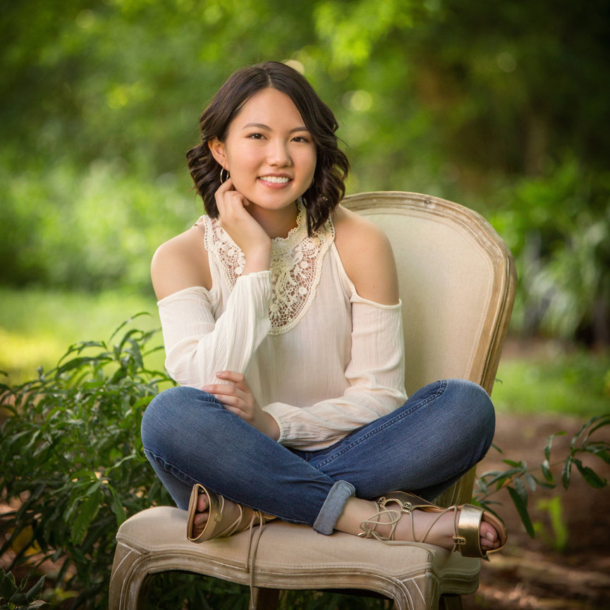 A person smiling while sitting on a chair outdoors, wearing a white top and jeans, surrounded by greenery.