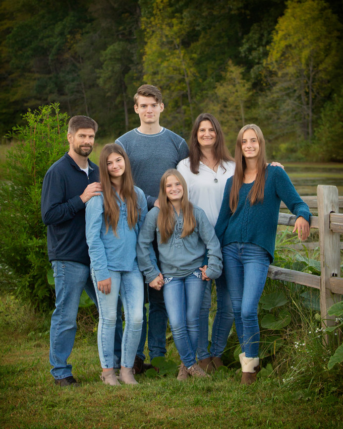 Family posing outdoors in casual outfits, standing by a wooden fence with greenery and trees in the background.