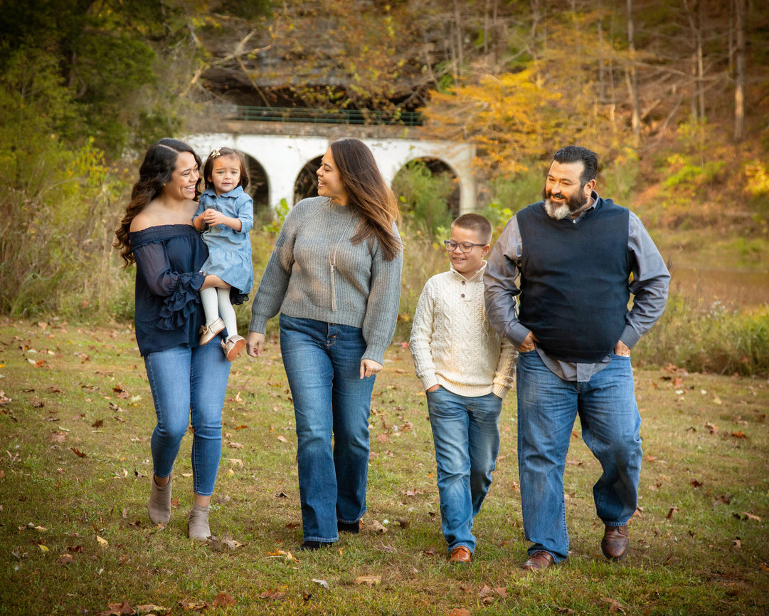 A family of five walks together in a park with autumn foliage and a stone bridge in the background.