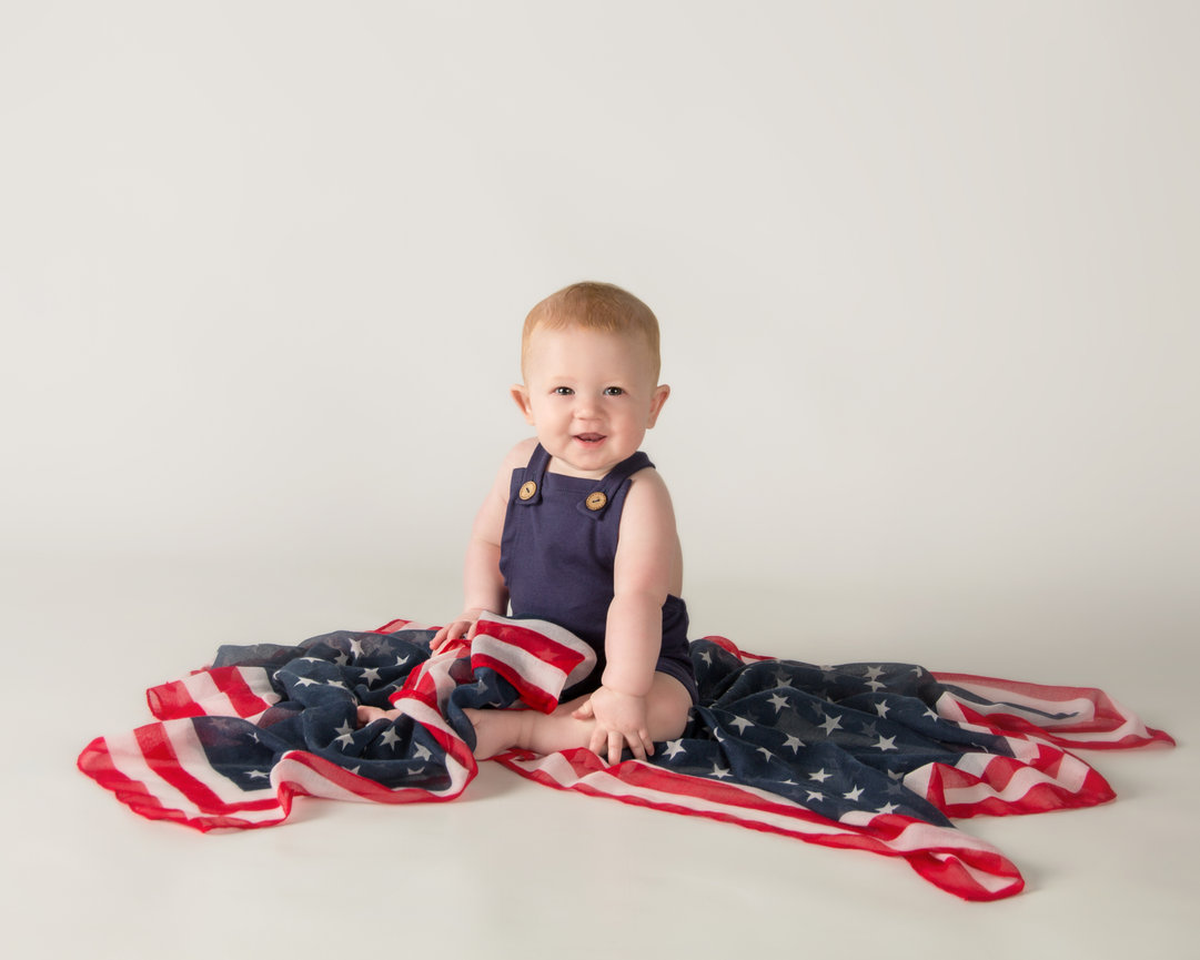 Baby sitting on a draped American flag, wearing a dark blue sleeveless outfit, against a plain background.