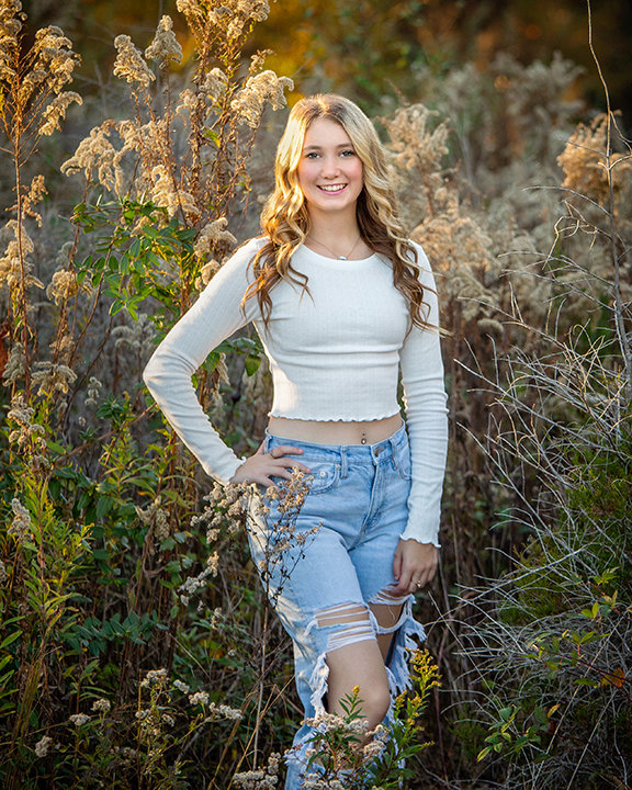 A person in a white long-sleeve top and ripped jeans stands in a sunlit field of tall grasses and plants.