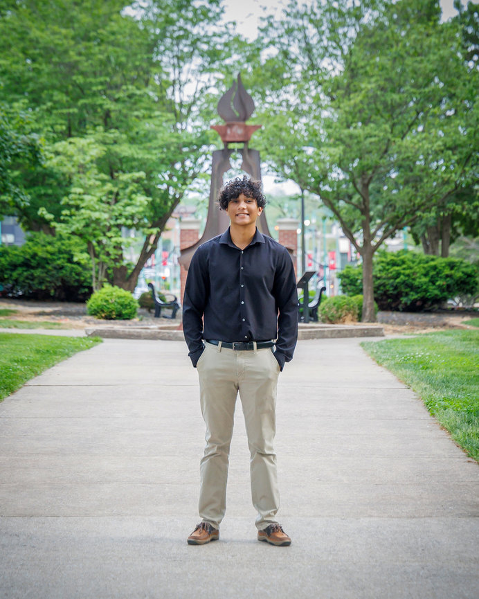 Young man in a black shirt and khaki pants standing on a pathway with trees and a monument in the background.