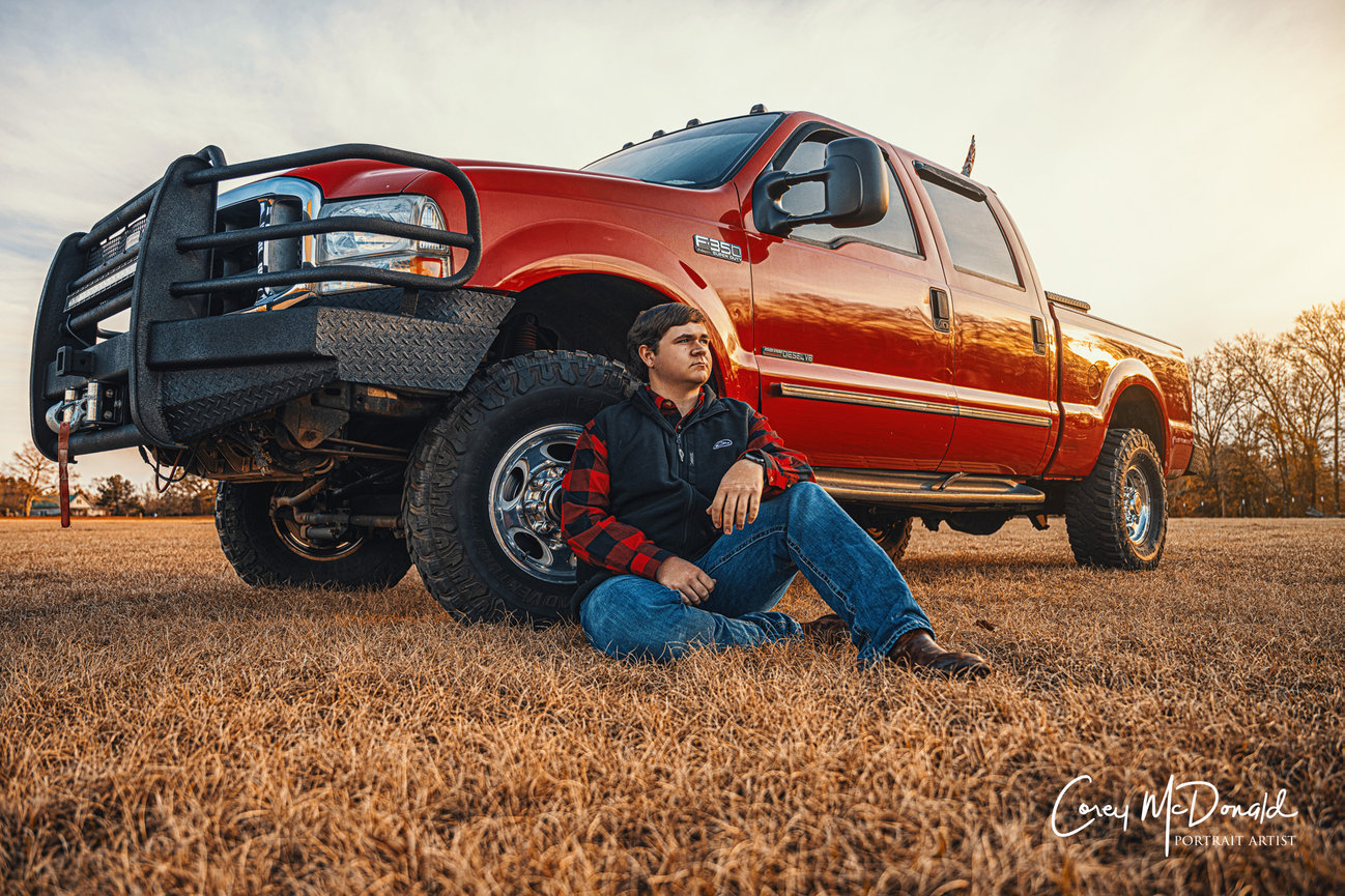 Man sitting on grass in front of a large red truck during sunset, wearing a black and red jacket and jeans.