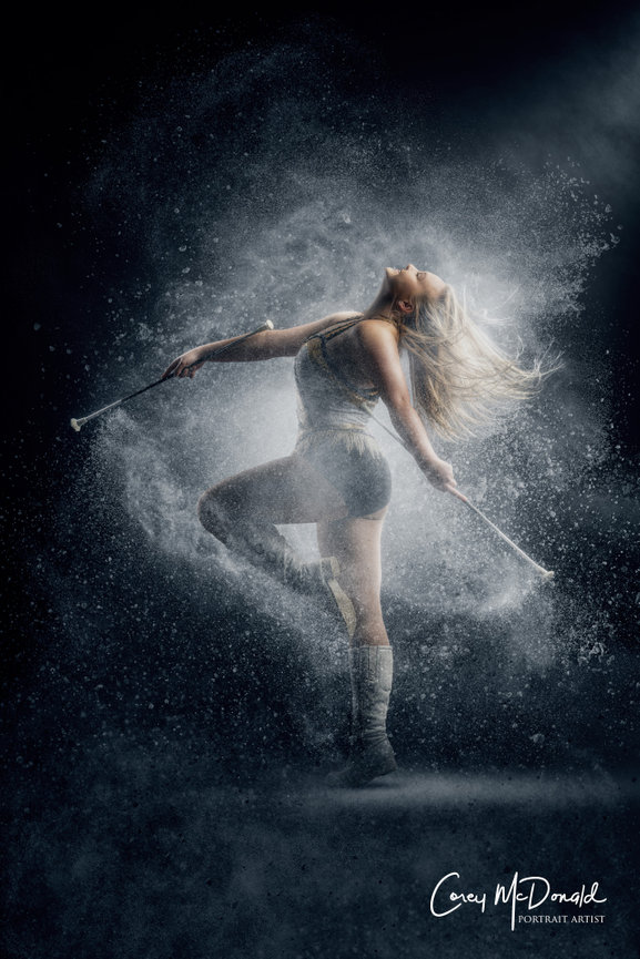 Dancer leaping with batons, surrounded by swirling powder on a dark background.