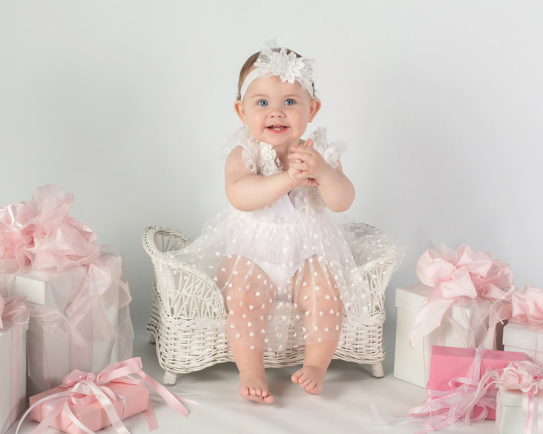 Smiling baby in a white dress and headband, sitting on a wicker chair, surrounded by pink gift boxes with ribbons.