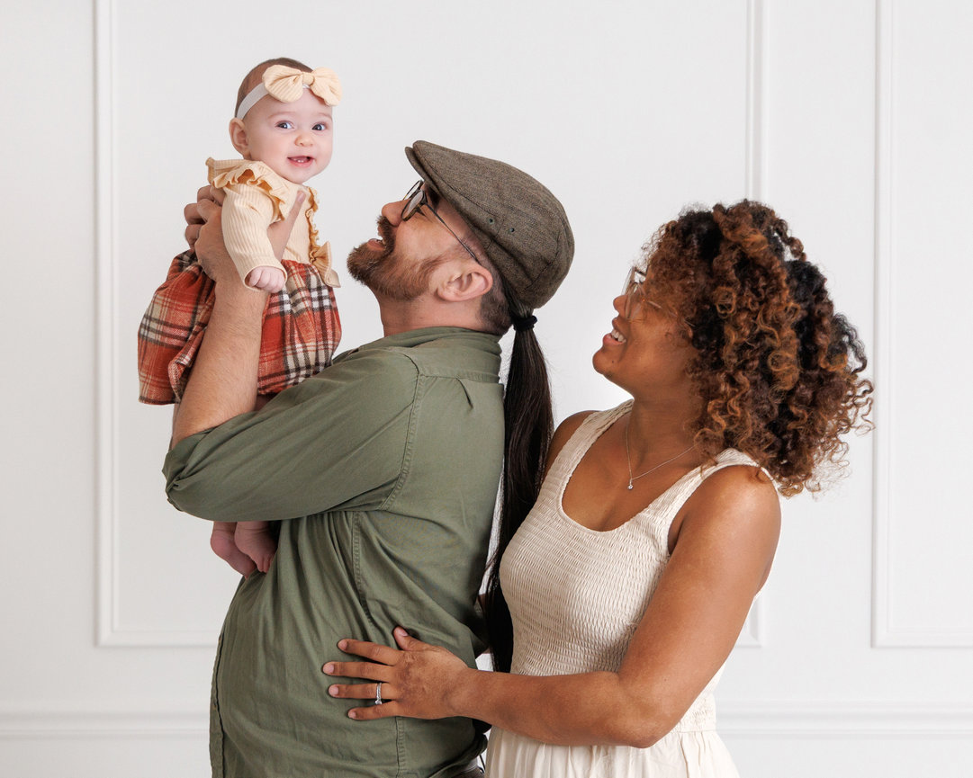 A couple smiling and holding a baby. The dad lifts the baby who wears a bow headband. Everyone looks joyful.
