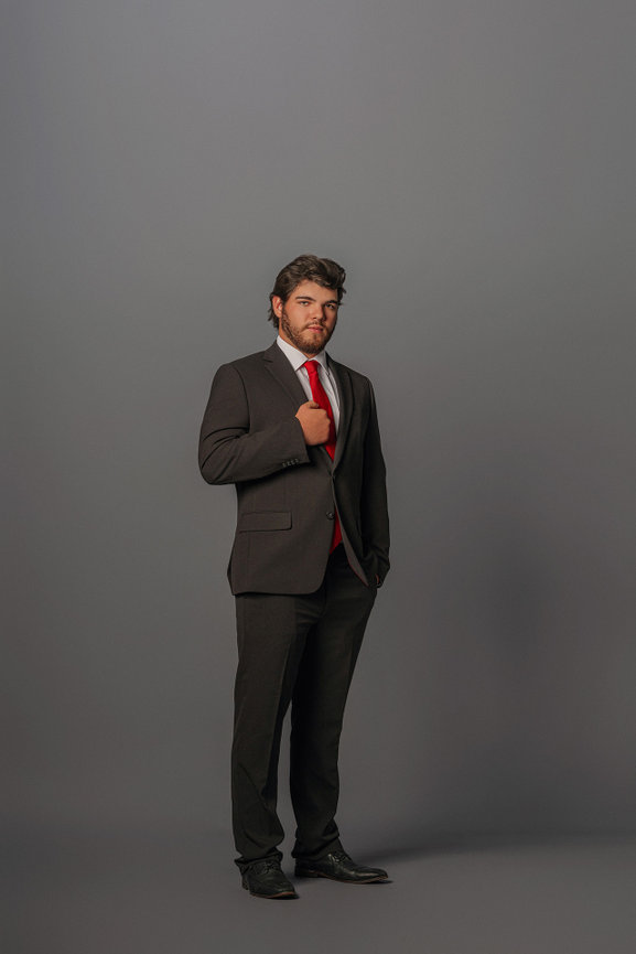 Man in a dark suit and red tie stands confidently against a gray background.