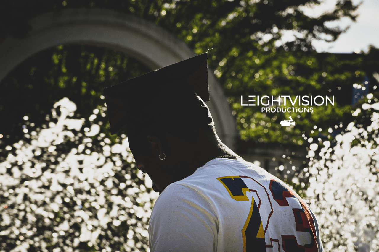A man in a graduation cap and t-shirt stands proudly in front of a fountain, celebrating his academic achievement.