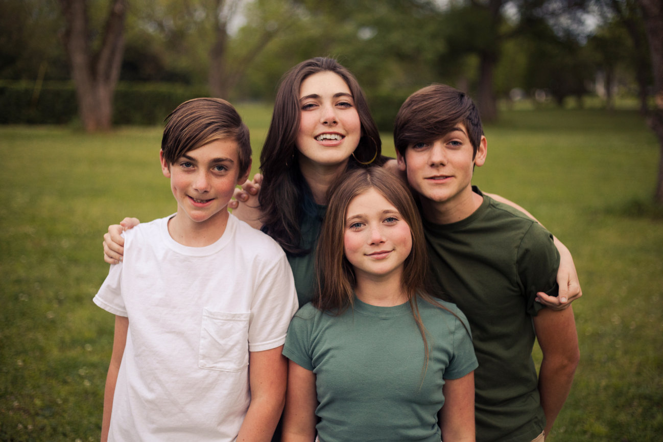Four siblings posing together outdoors with a blurred park background.