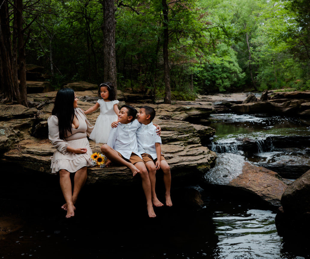 A woman and three children sit on rocks by a forest stream, surrounded by lush greenery.