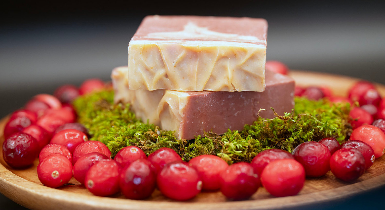 Artisanal soap bars on moss, surrounded by cranberries on a wooden plate