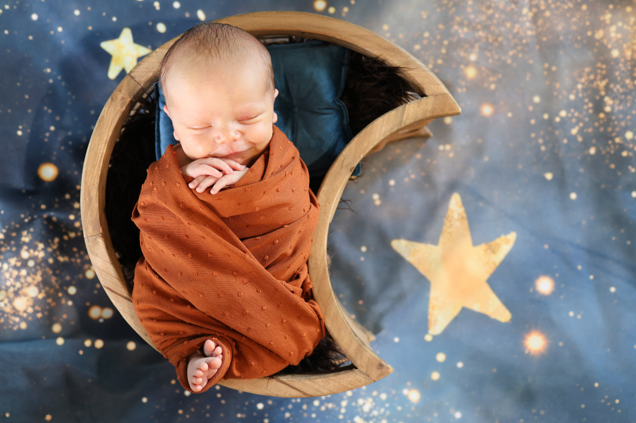 Newborn baby wrapped in rust-colored fabric on a crescent moon prop with stars