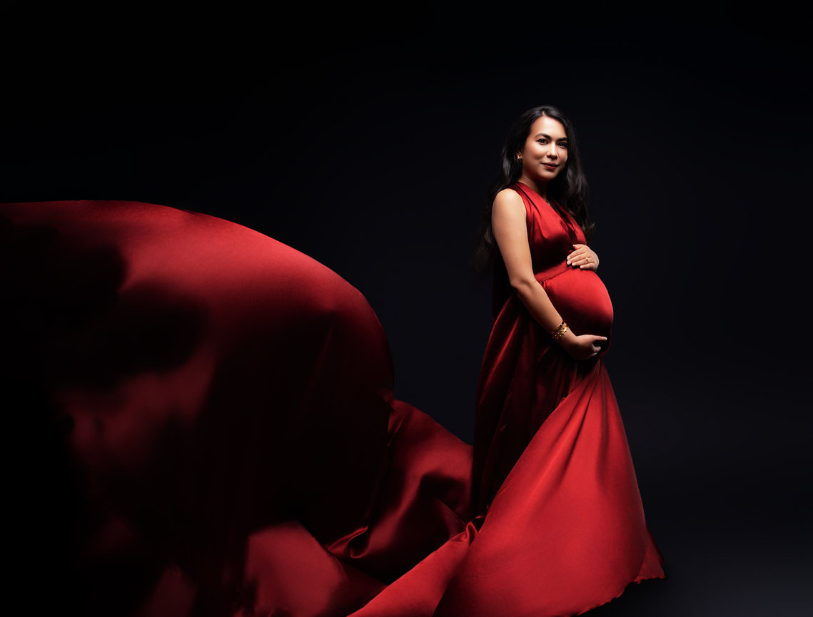 Elegant maternity portrait of pregnant woman in flowing red gown, taken in a high-end photography studio with dramatic lighting