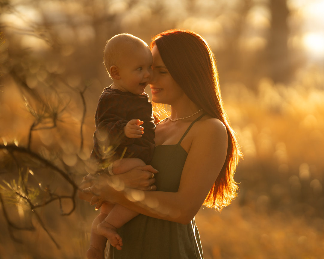 Mother holding baby in sunlit field, both smiling warmly, surrounded by soft-focus foliage and golden light.