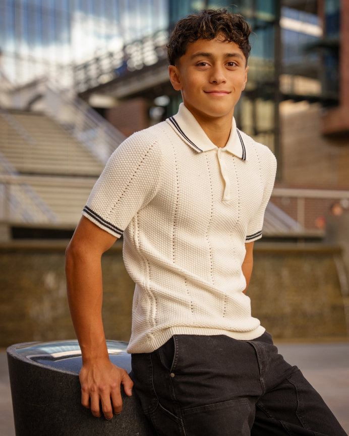 A person in a white polo shirt and dark pants leans against a stone structure outdoors, with a modern building in the background.