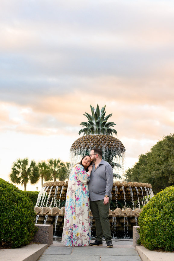 couple huddles close and man kisses woman on the head against a sunset sky and charlestons pineapple fountain