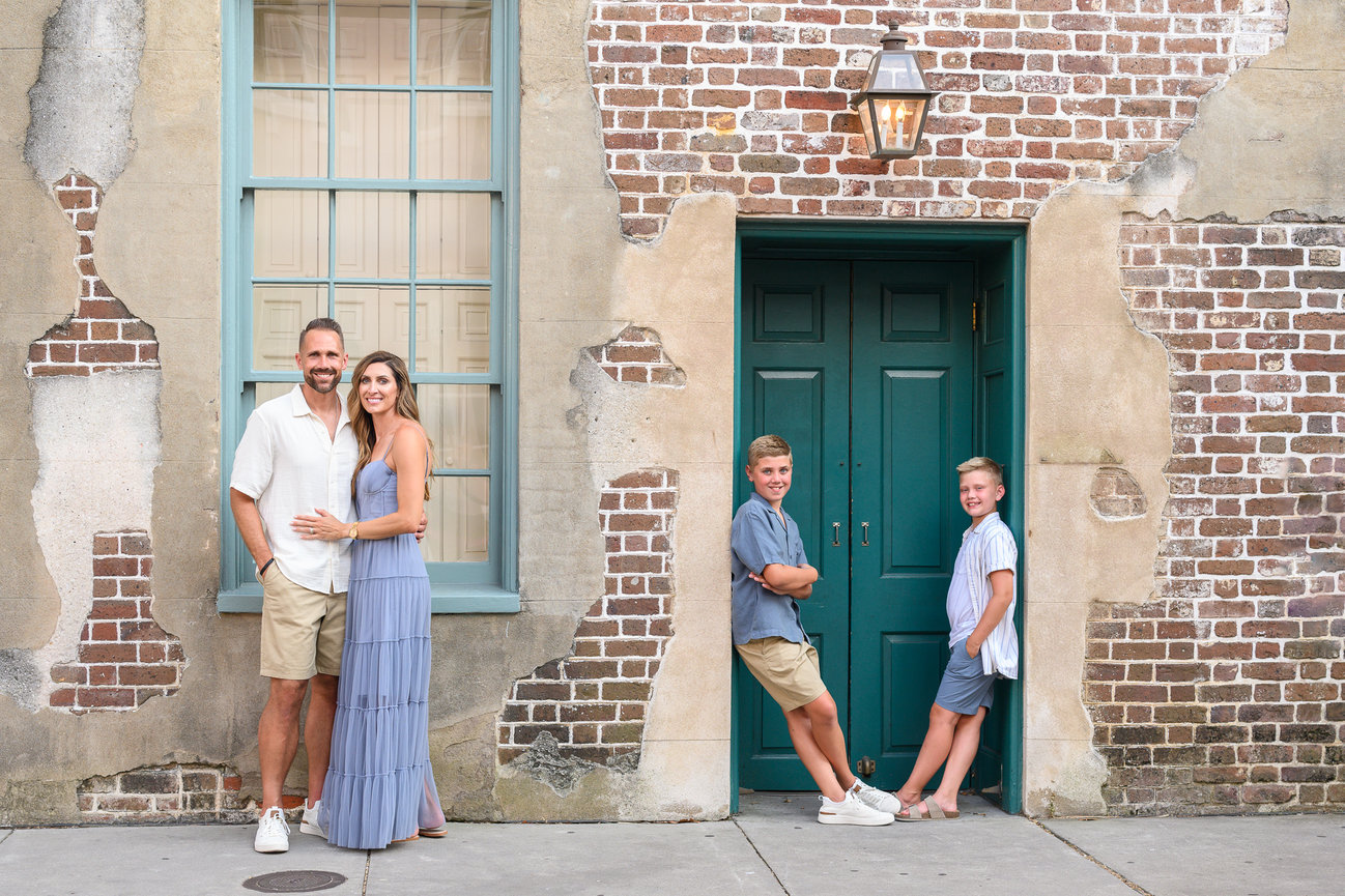 A family of four posing in front of a textured brick wall with two green doors and a lantern.