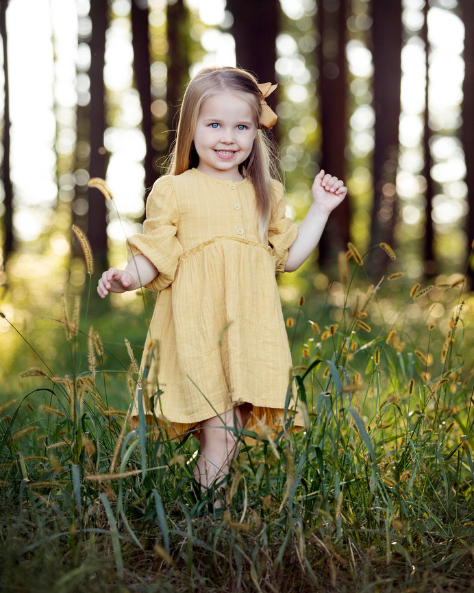 Smiling child in a yellow dress standing in a grassy forest clearing with sunlit trees behind her
