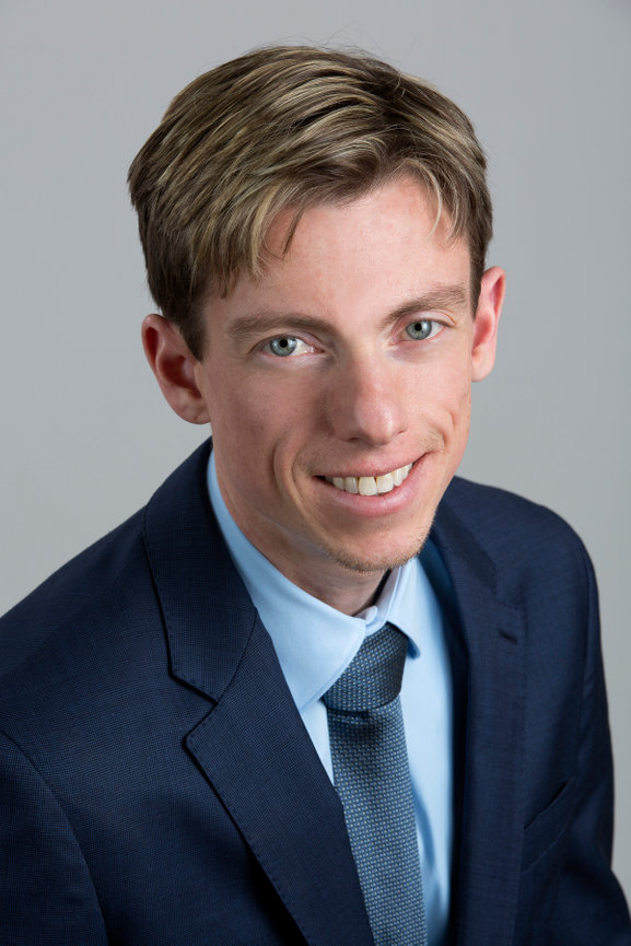 Headshot of a smiling young man in a suit and tie against a dark background