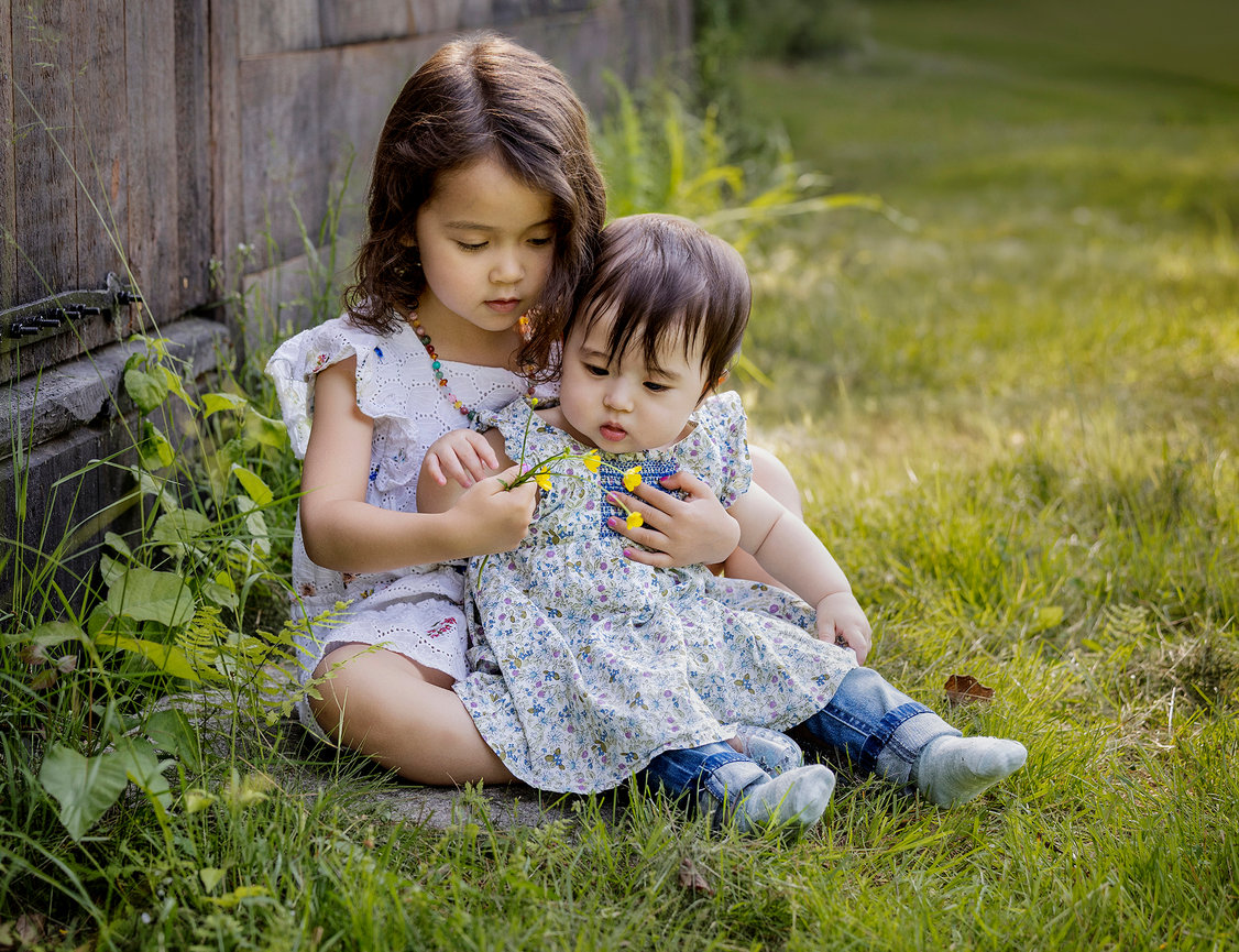 Two young children sitting on grass, with the older one showing a flower to the younger.