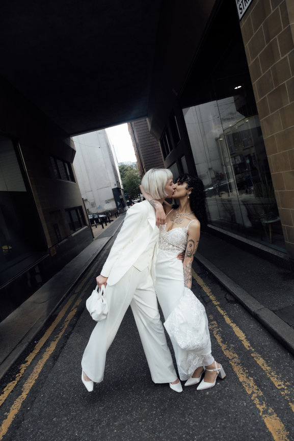 Editorial wedding photography of two brides sharing a joyful kiss on an empty Manchester city street, one bride leaning in with style while wearing a modern white wedding suit.