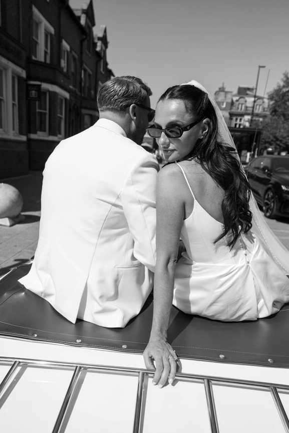 Black & White wedding photography outside The Terrace in Warrington. Bride and groom in white sit on a car, with the bride looking back, both wearing sunglasses.