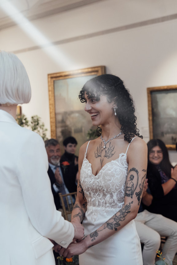A smiling, tattooed bride in a sunlight shaft during a same sex wedding ceremony at Manchester Art Gallery, captured in an editorial photography style.