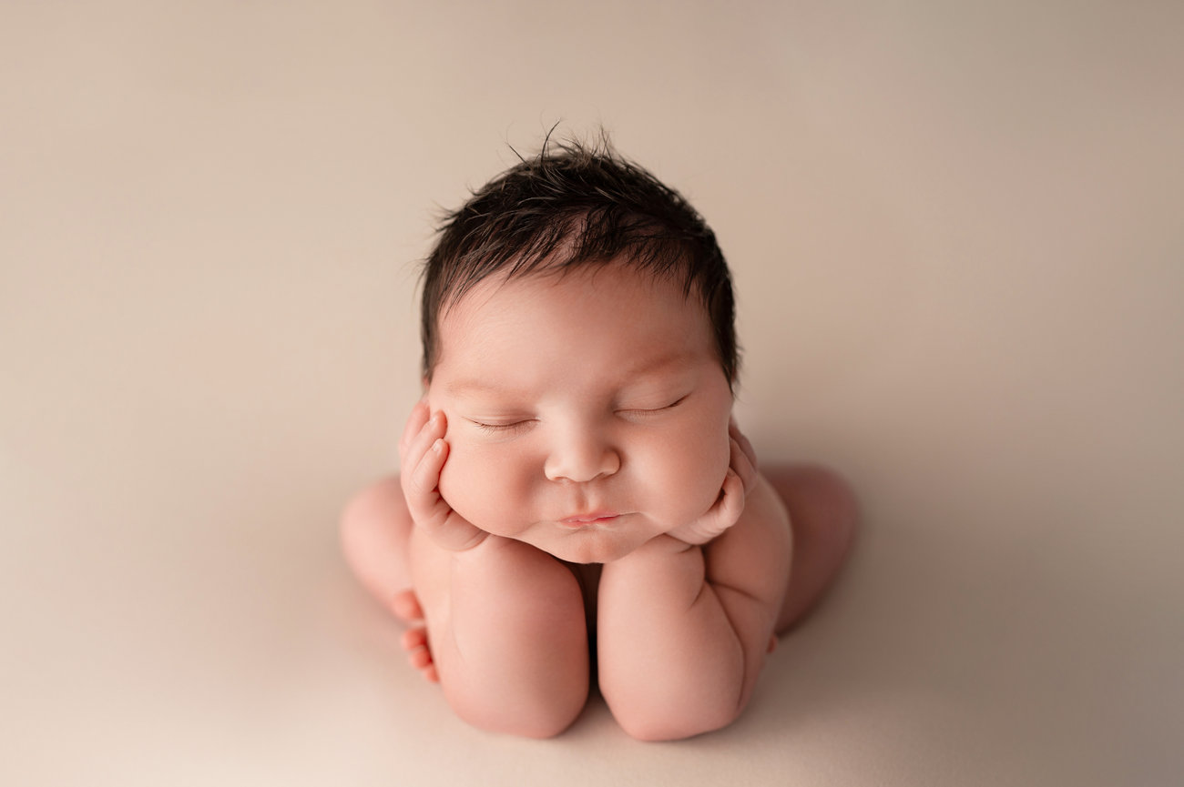 Peaceful newborn baby in a froggy pose with hands tucked under chin on solid tan background