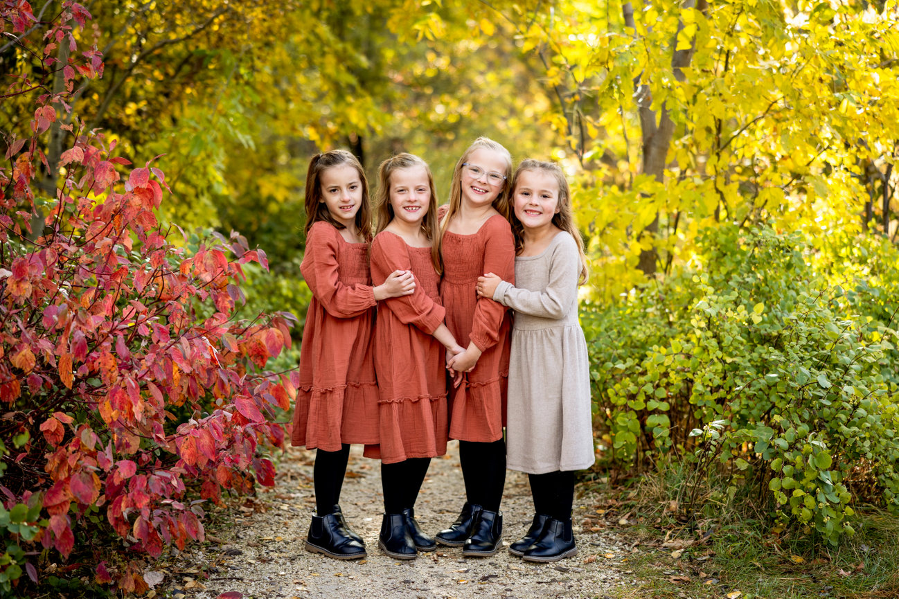 Four girls in autumn dresses stand together on a path surrounded by colorful fall foliage.