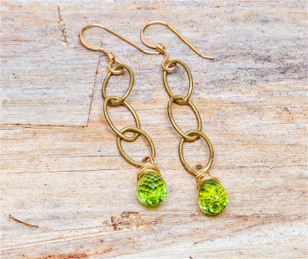 Pair of gold dangle earrings with three interlocking oval links, each featuring a faceted teardrop-shaped green gemstone wrapped in gold wire, displayed on a rustic wooden background.