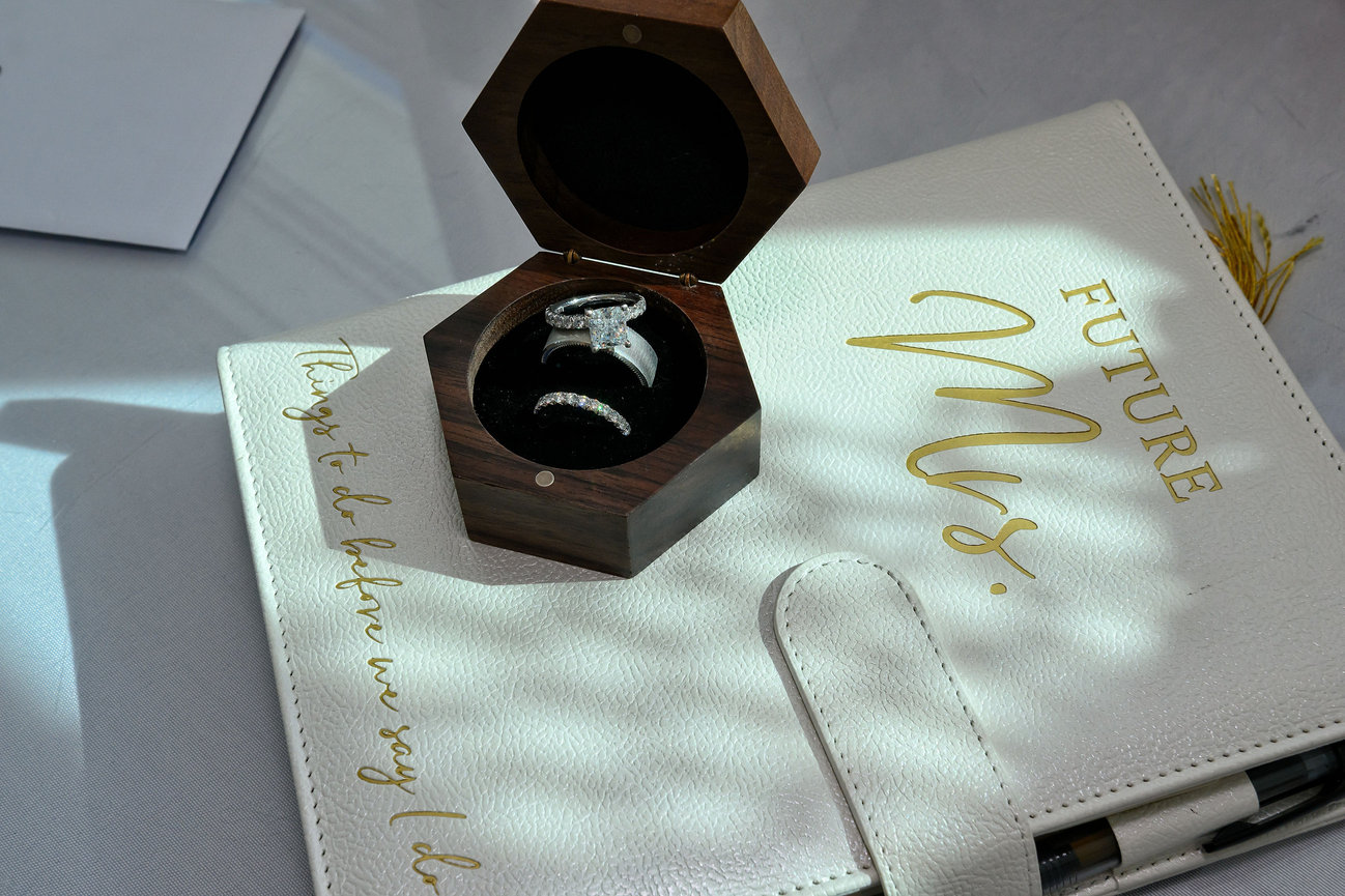 Open wooden ring box containing a diamond engagement ring and wedding band, resting on a white leather planner with gold lettering that reads 'Future Mrs.' and 'Things to do before we say I do.