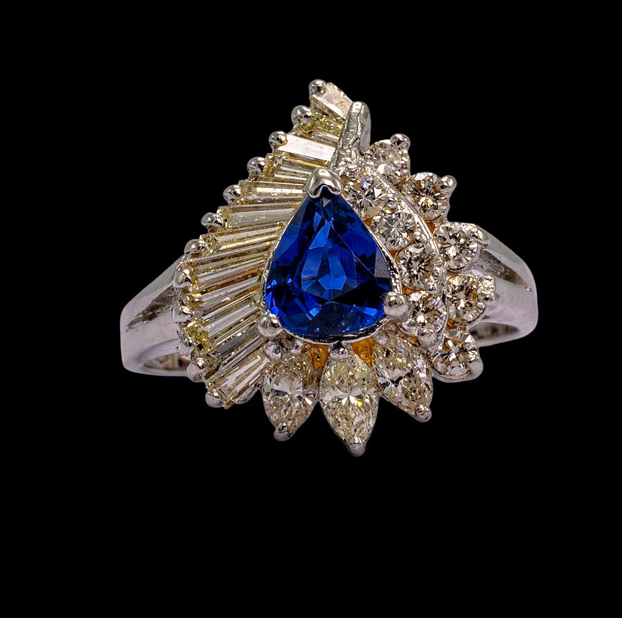Platinum ring featuring a pear-shaped blue sapphire centerpiece surrounded by a fan of tapered baguette and round brilliant-cut diamonds, accented with marquise-cut yellow diamonds, set against a black background.