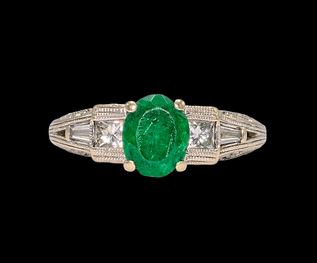 latinum and gold ring featuring an oval-cut emerald centerpiece flanked by two square-cut diamonds and tapered baguette diamonds, set against a black background.