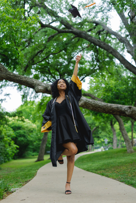 A graduate in a black gown joyfully throws her cap into the air while walking along a tree-lined path at Prairie Creek Park in Richardson, TX