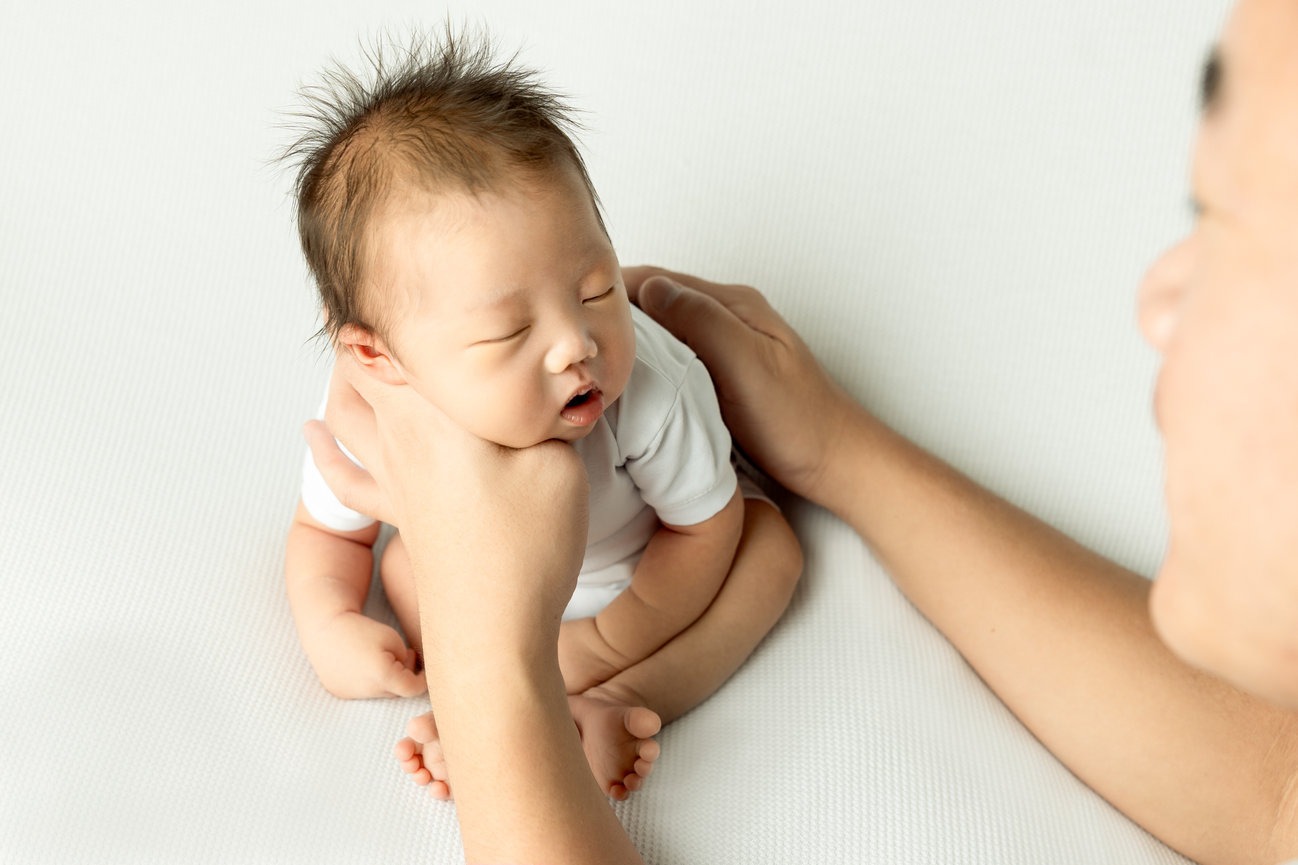 A sleeping baby with tousled hair held gently by an adult's hands on a white surface.