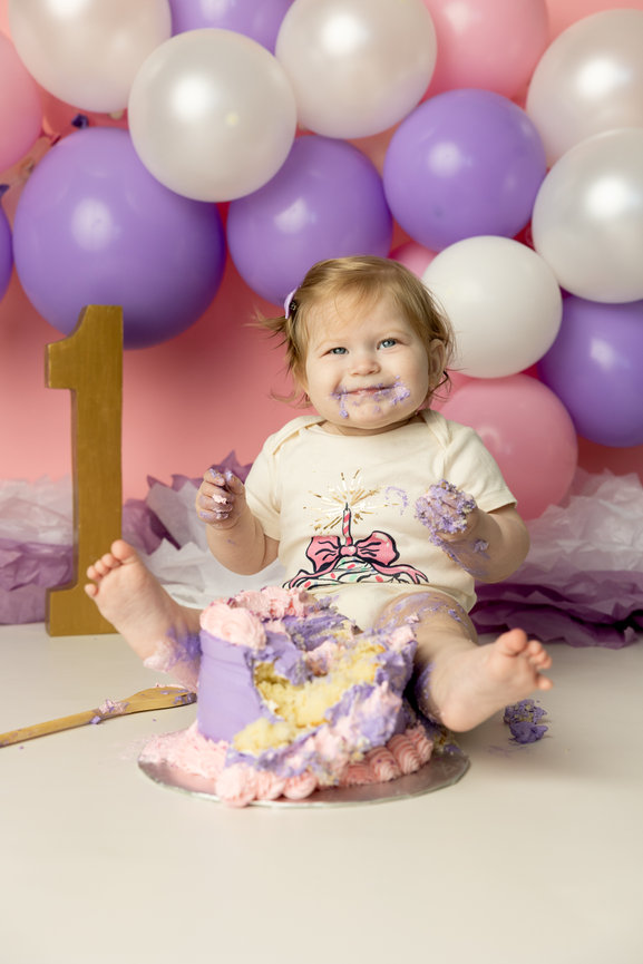 Baby with cake on face and hands, sitting beside a purple and pink cake, surrounded by balloons and a gold number one.
